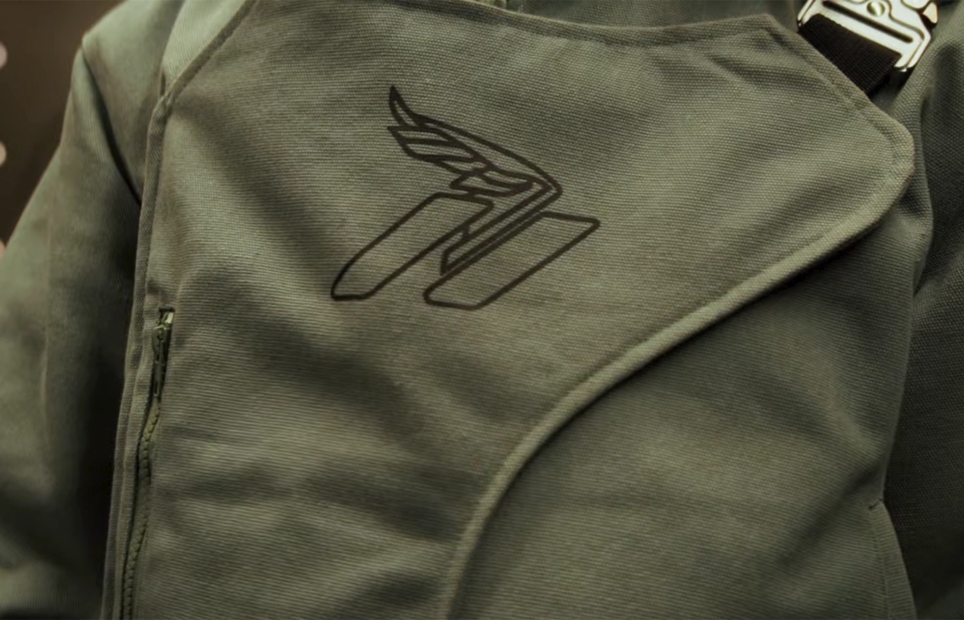 Close-up of an olive green jacket with an embroidered wing logo on the chest and a metal buckle strap
