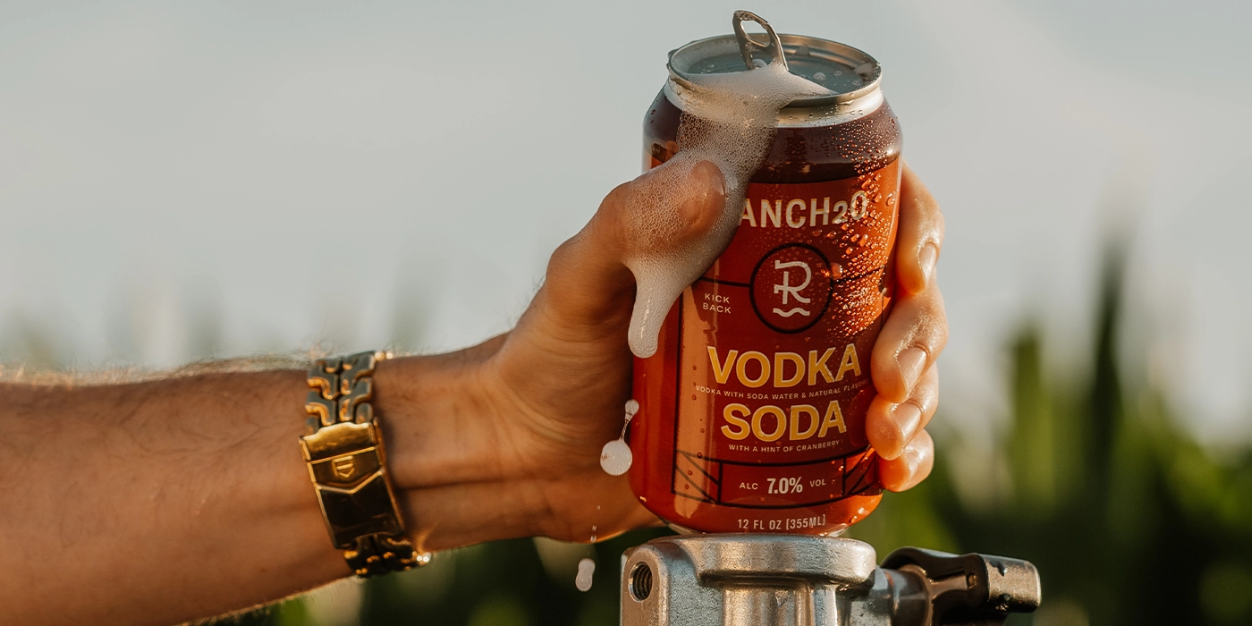 Hand wearing a gold bracelet gripping a freshly opened, foaming can of Ranch2O Vodka Soda outdoors.